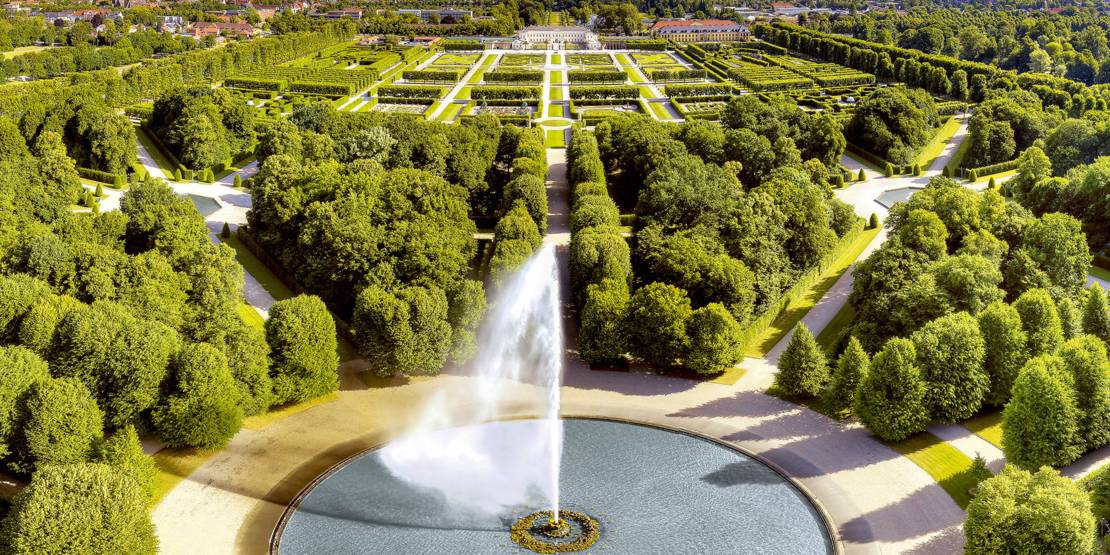 Hier sieht man eine Drohnenaufnahme der großen Fontäne in den Herrenhäuser Gärten mit Blick auf das Schloss Herrnhausen