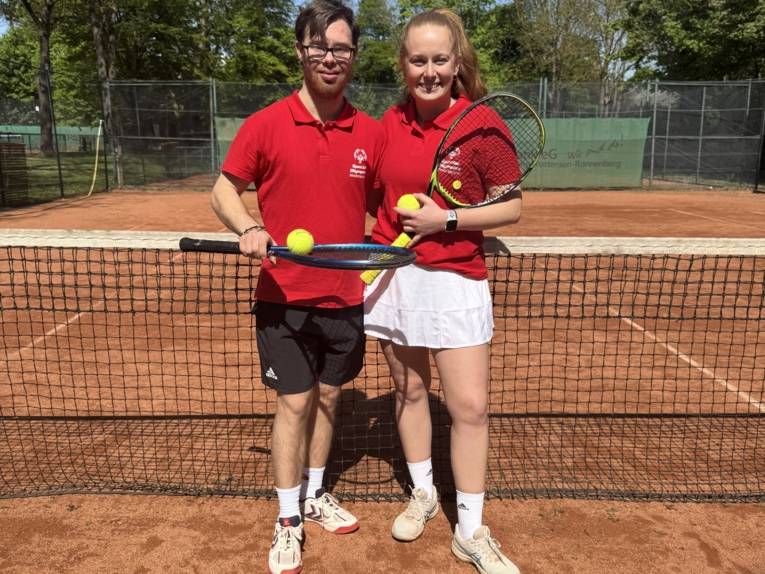 Zwei Tennis-Spielerim roten Teamshirt posieren mit Schlägern und Bällen vor dem Netz auf Sandplatz.