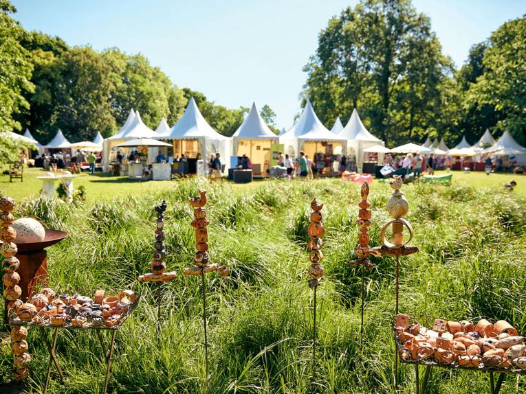 Kunsthandwerkermarkt im Grünen mit weißen Pagodenzelten, dekorativen Keramiken im Vordergrund und Besuchern zwischen den Ständen.